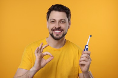 Photo of Happy man holding electric toothbrush and showing ok gesture on yellow background