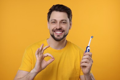 Happy man holding electric toothbrush and showing ok gesture on yellow background