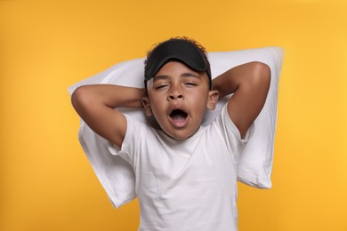 Photo of Boy with pillow and sleep mask yawning on yellow background. Insomnia problem