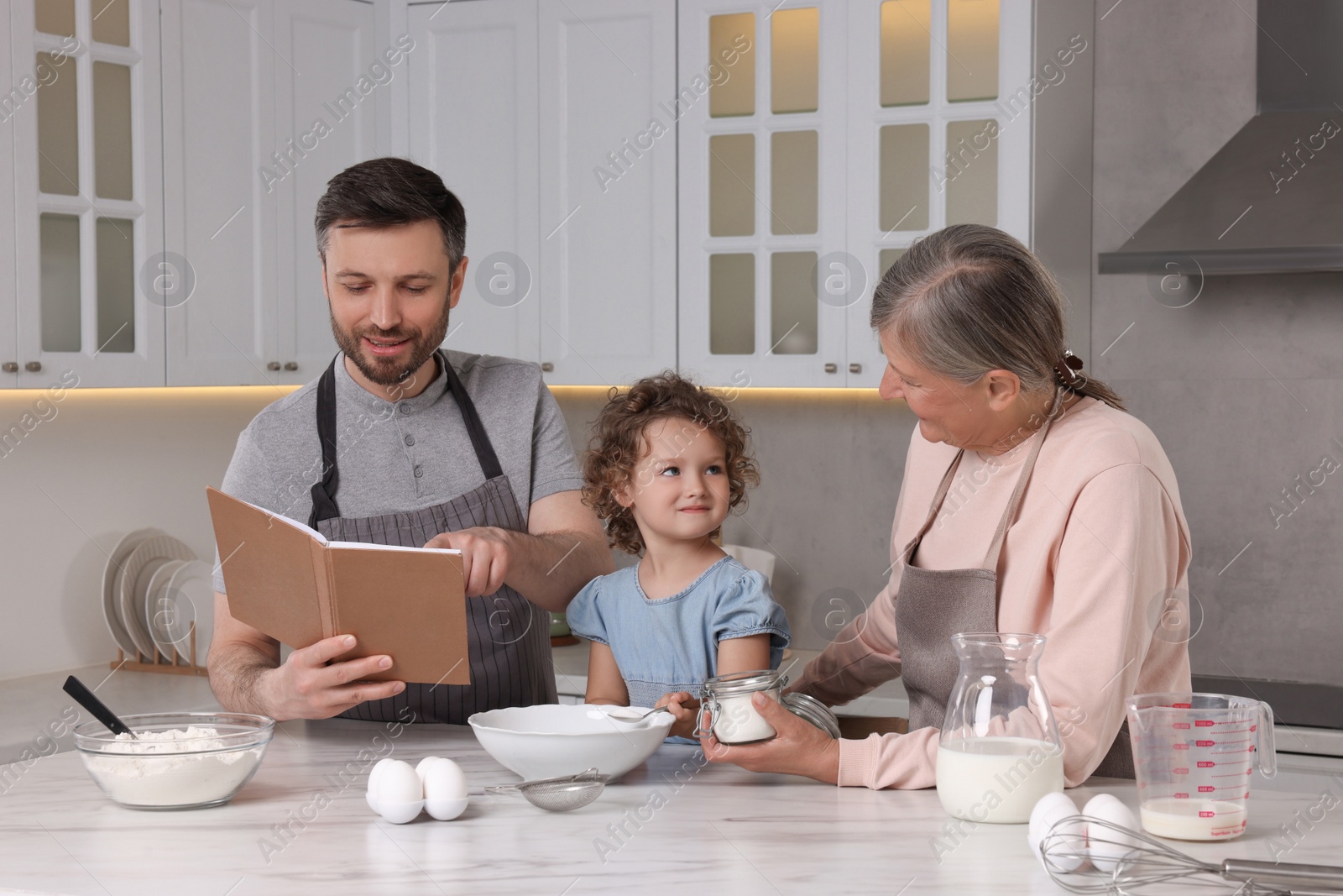 Photo of Cute little girl with her father and grandmother cooking by recipe book in kitchen