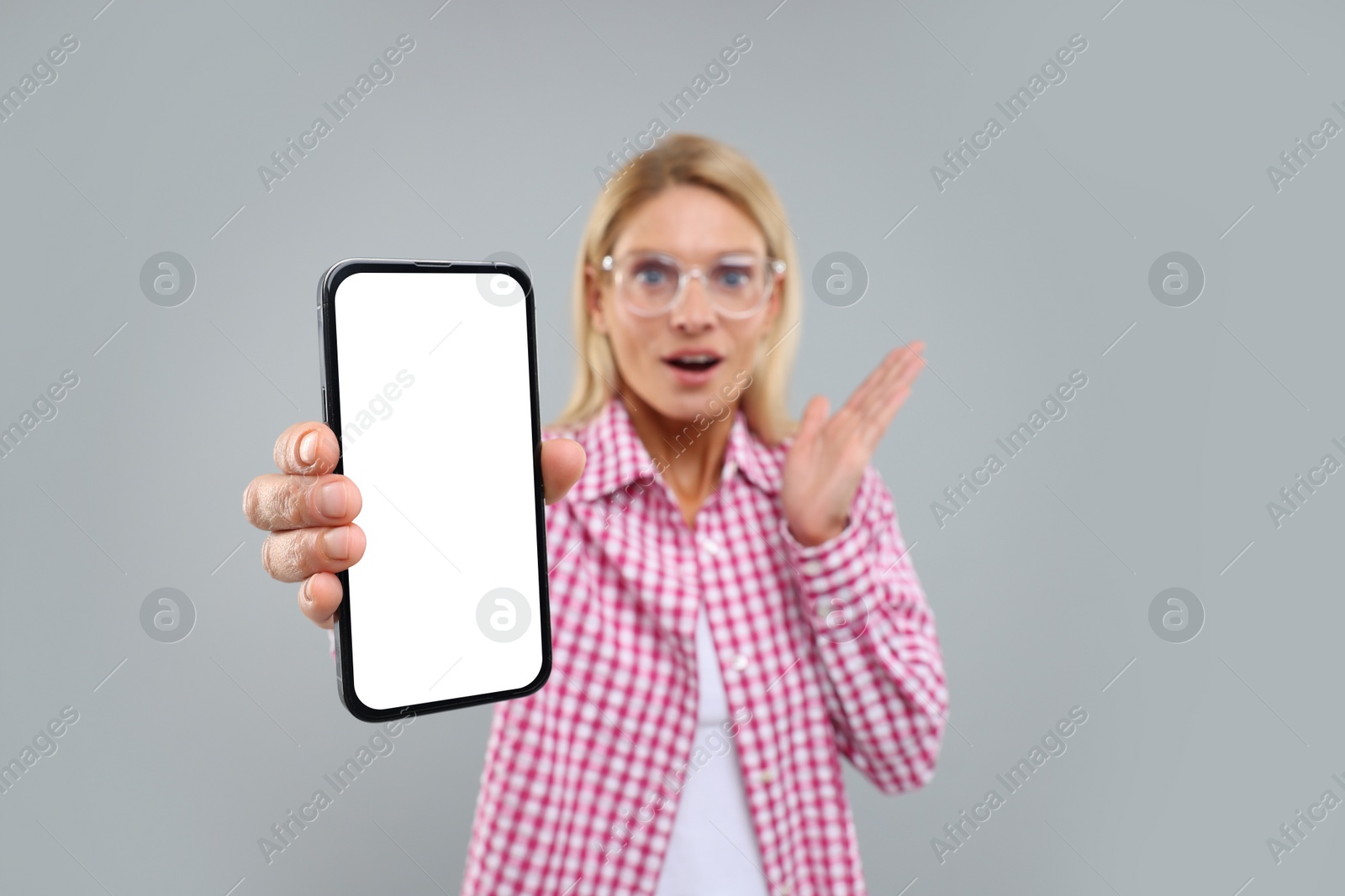 Photo of Surprised woman holding smartphone with blank screen on grey background, selective focus