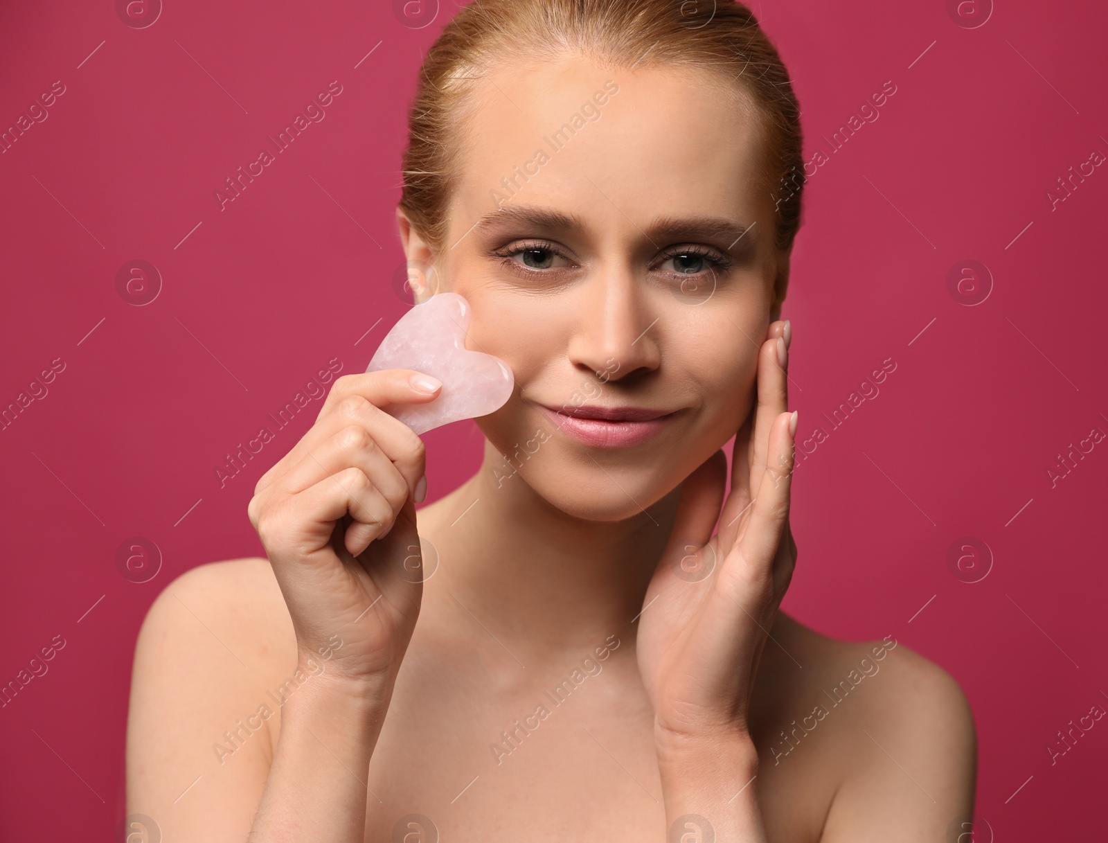 Photo of Beautiful young woman doing facial massage with gua sha tool on pink background, closeup