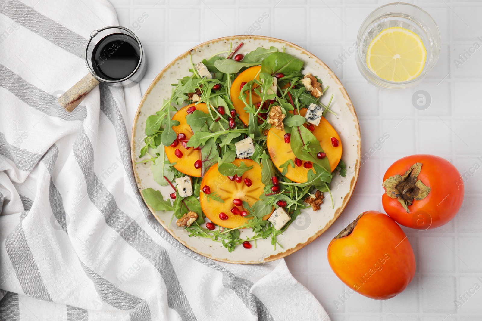 Photo of Tasty salad with persimmon, blue cheese, pomegranate and walnuts served on white tiled table, flat lay