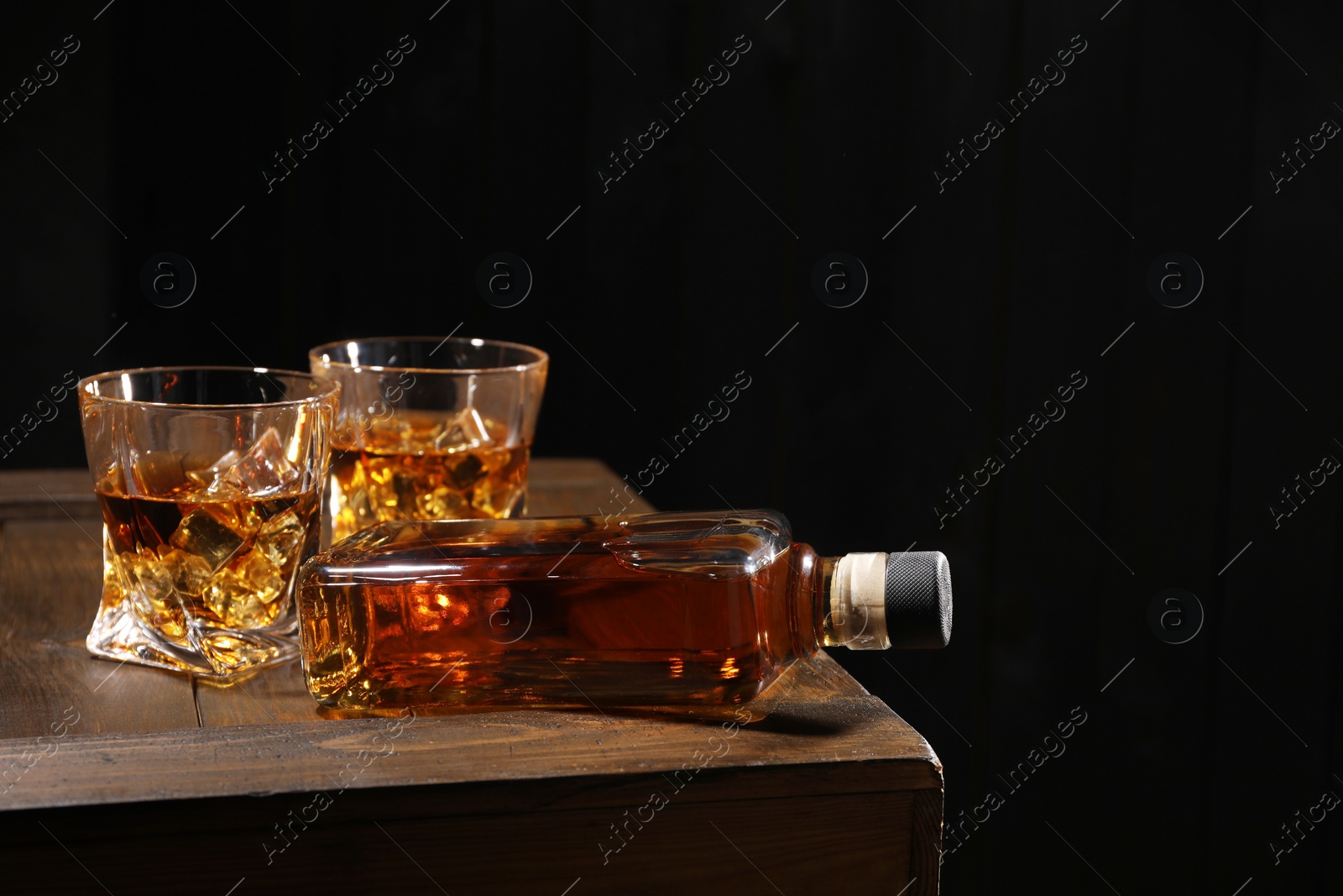Photo of Whiskey with ice cubes in glasses and bottle on wooden crate against black background, closeup. Space for text
