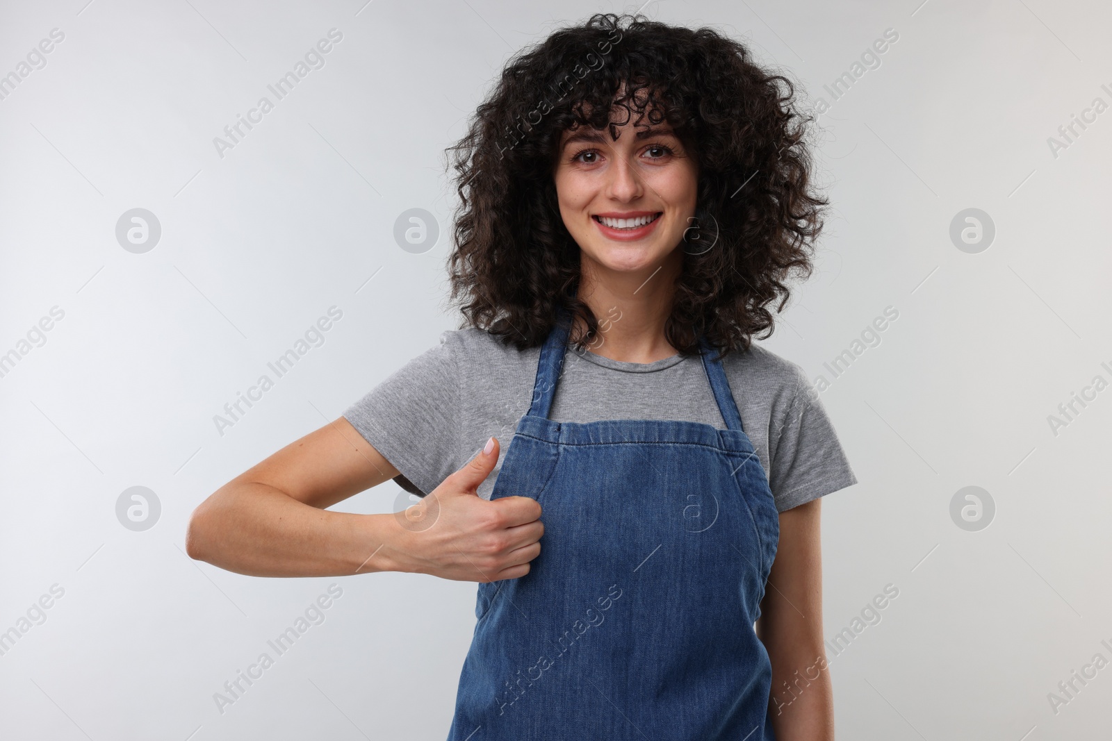 Photo of Happy woman wearing kitchen apron and showing thumbs up on light grey background. Mockup for design