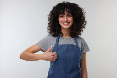 Happy woman wearing kitchen apron and showing thumbs up on light grey background. Mockup for design