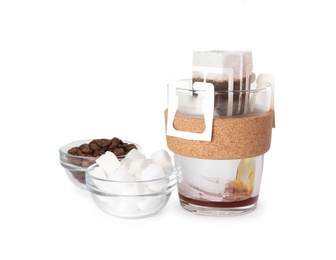 Photo of Pouring hot water into glass cup with drip coffee bag, near beans and sugar cubes isolated on white