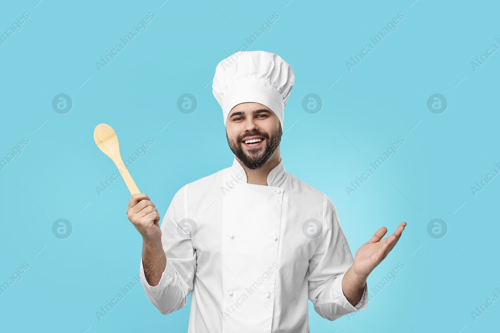 Photo of Happy young chef in uniform holding wooden spoon on light blue background