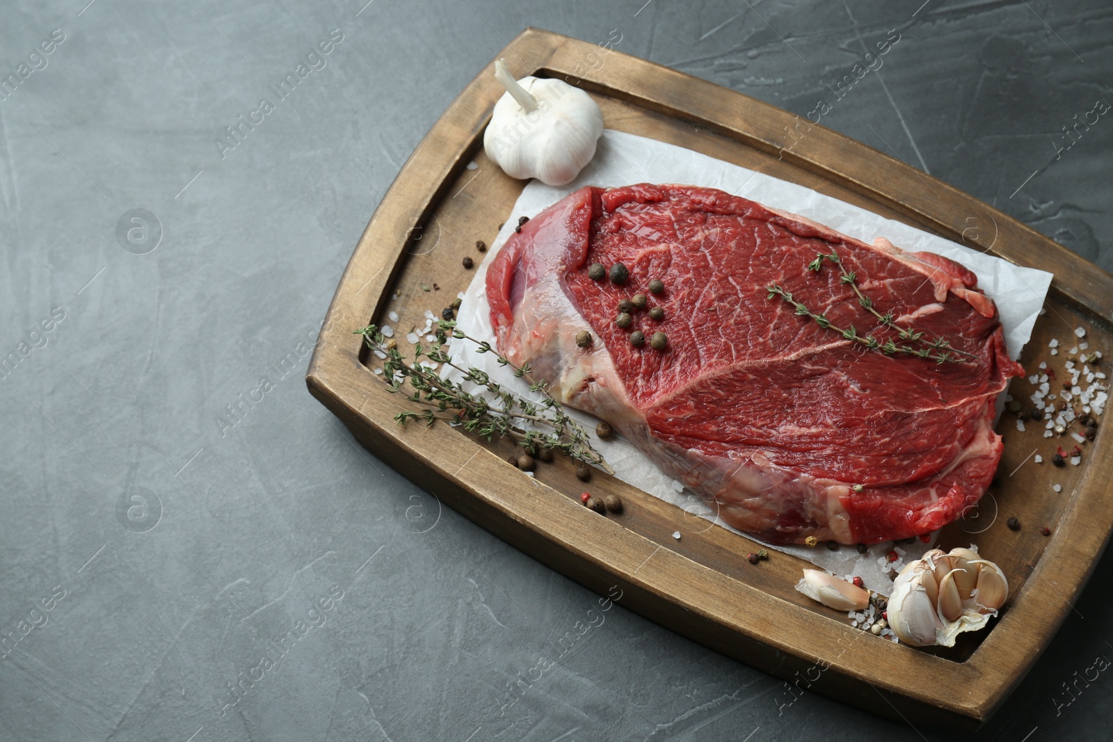 Photo of Wooden tray with piece of raw meat, garlic and thyme on grey table, above view. Space for text