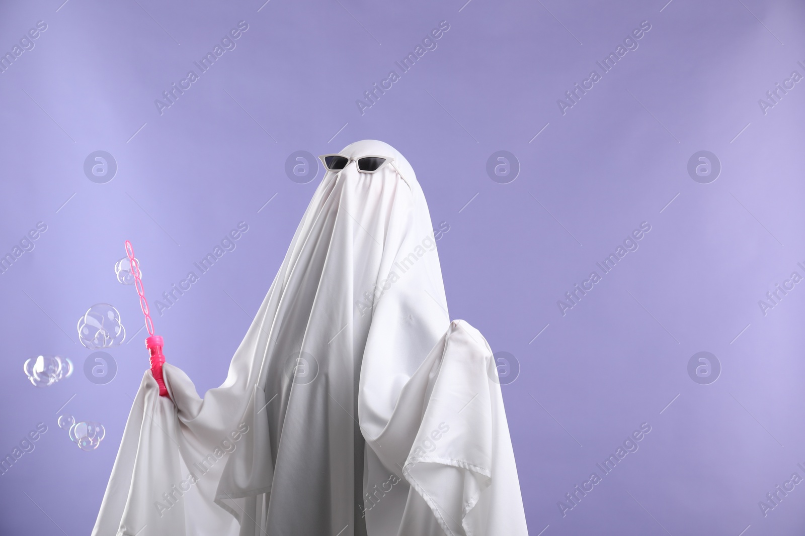 Photo of Funny ghost. Person in white sheet, sunglasses and soap bubbles on violet background, space for text