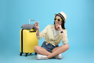 Photo of Happy female tourist with toy plane, suitcase and travel pillow on light blue background