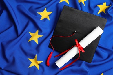 Photo of Black graduation cap and diploma on flag of European Union, flat lay. Space for text