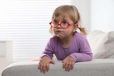 Cute little girl in glasses on sofa at home. Space for text