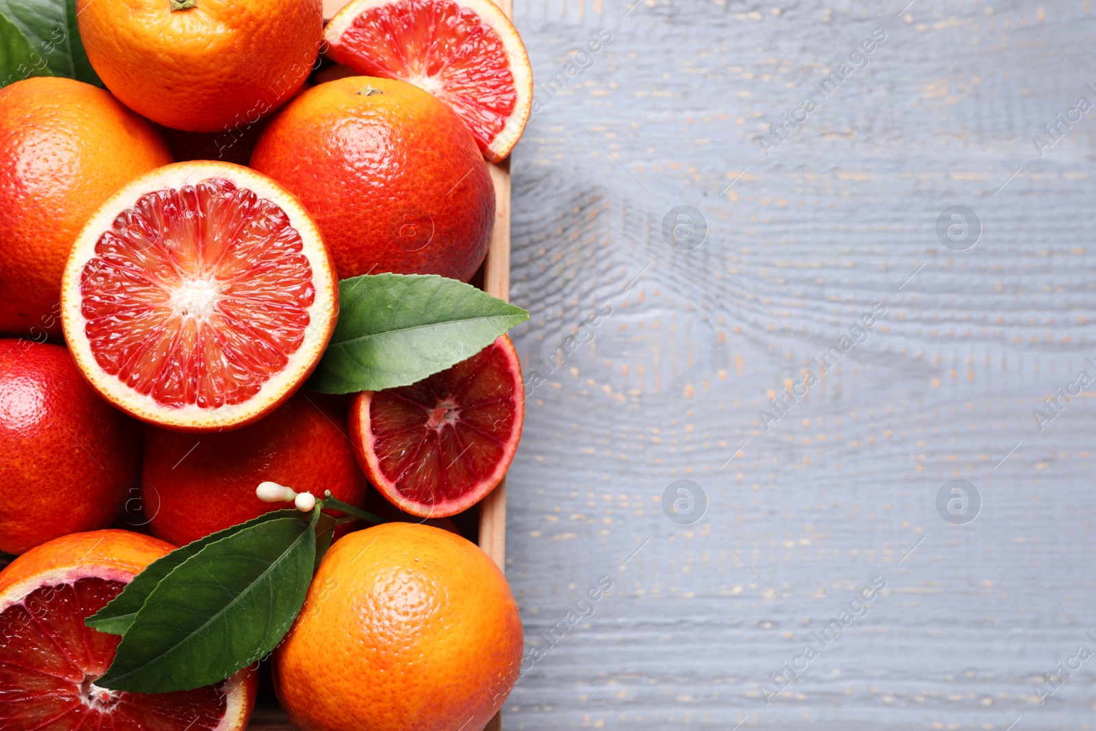 Photo of Crate of ripe red oranges and green leaves on grey wooden table, top view. Space for text