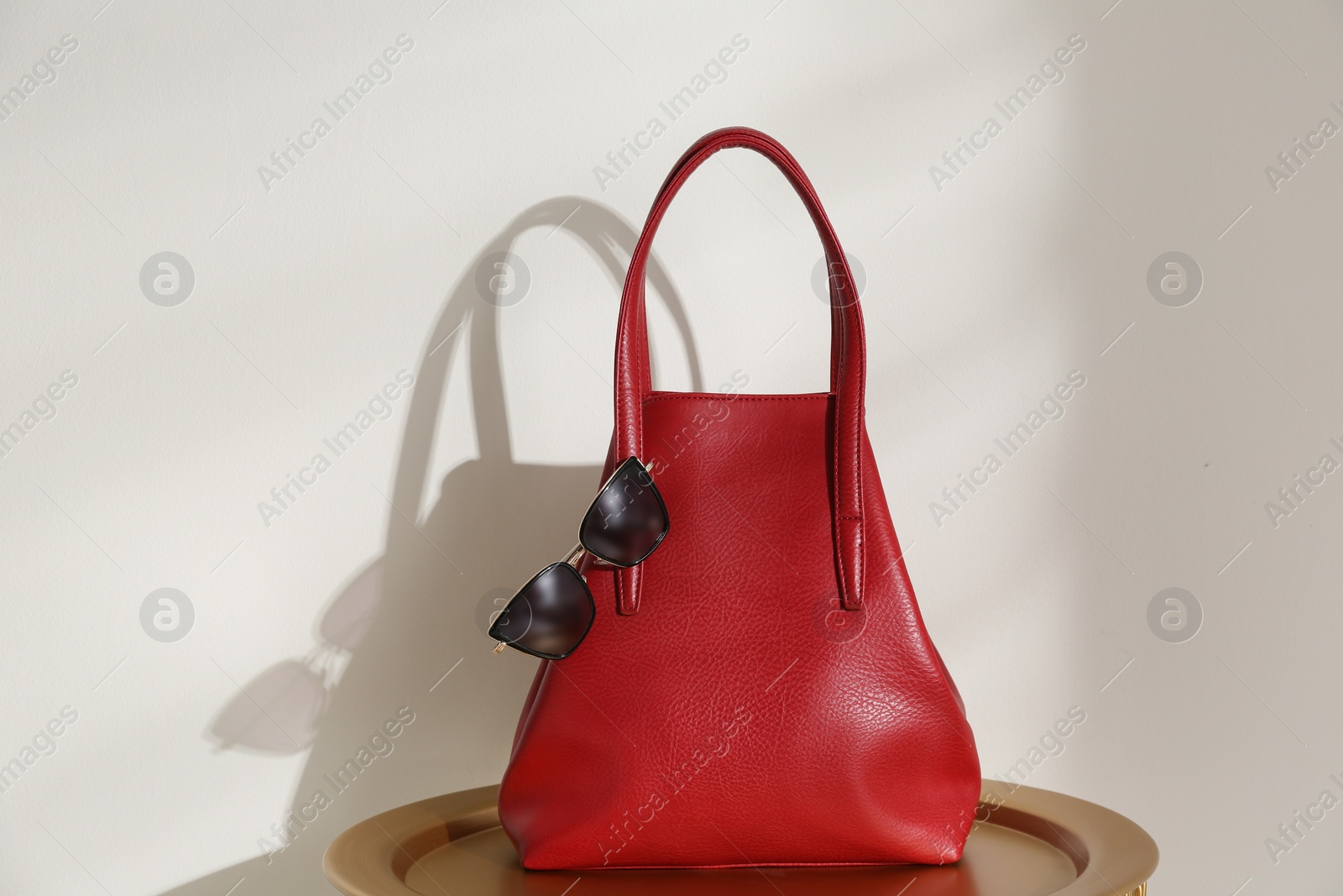 Photo of Stylish red woman's bag and sunglasses on table near light wall