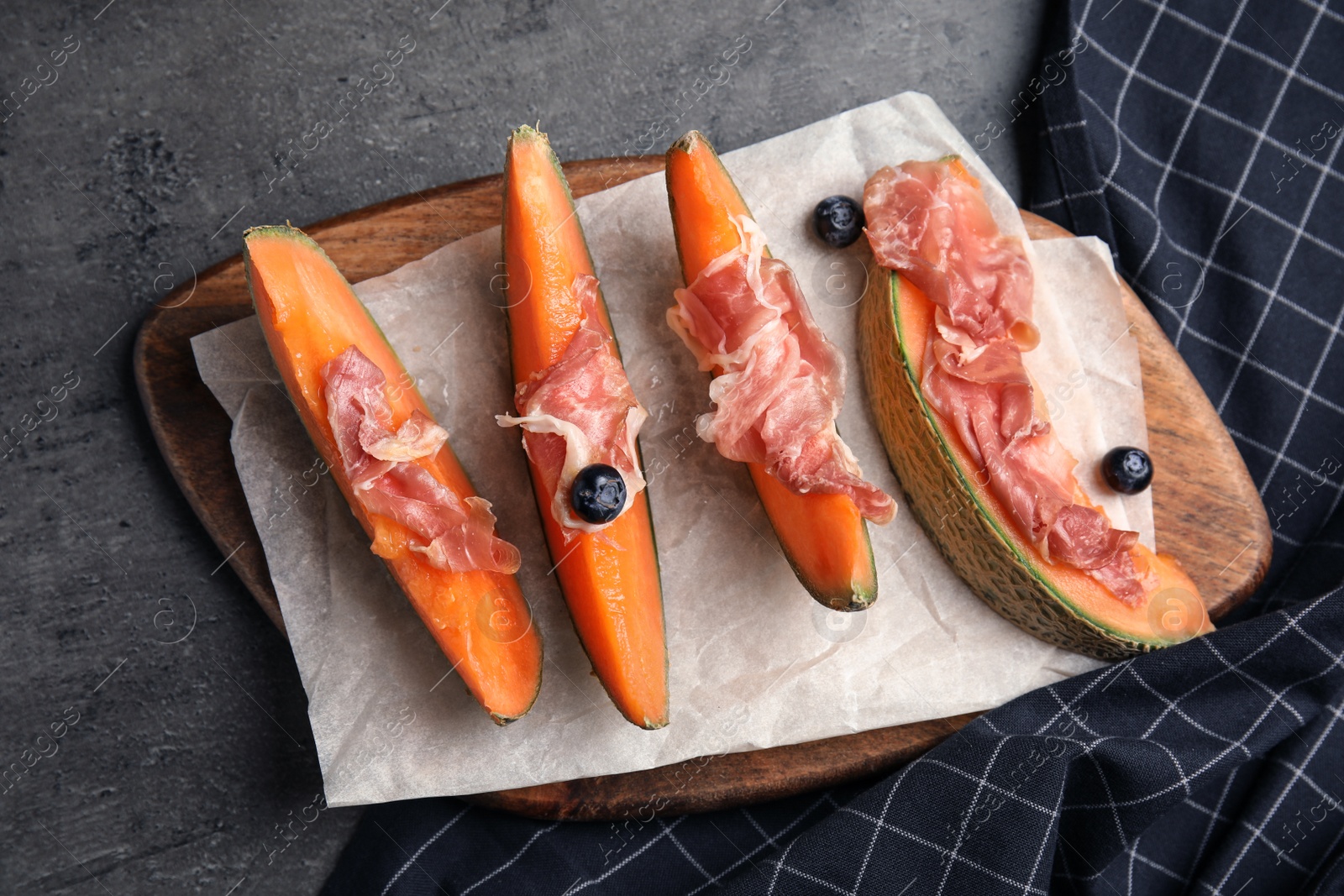 Photo of Wooden board with melon, prosciutto and blueberries on grey table, above view