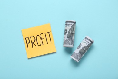 Photo of Sticky note with word Profit and rolled banknotes on turquoise background, flat lay