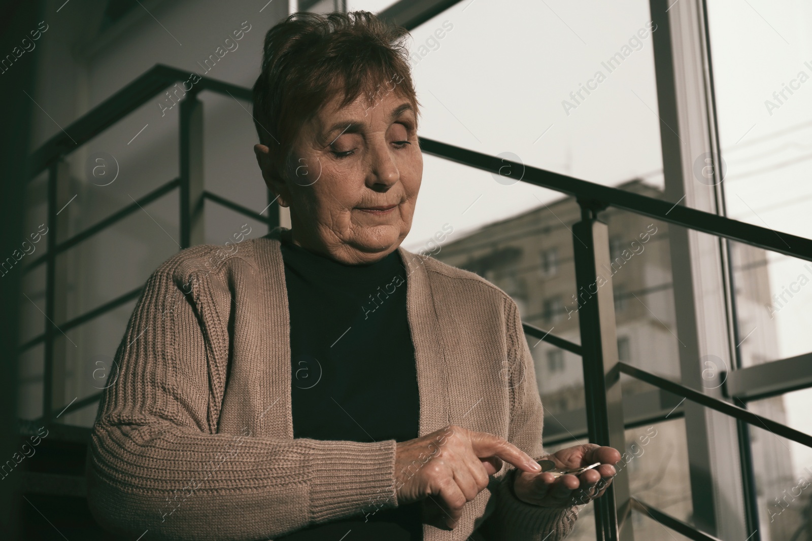 Photo of Poor senior woman with coins sitting on stairs indoors