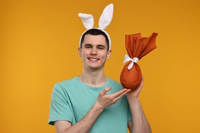 Photo of Easter celebration. Handsome young man with bunny ears holding wrapped gift on orange background