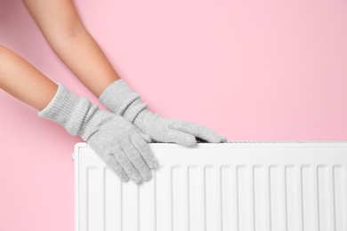 Photo of Woman in gloves warming hands on heating radiator near color wall