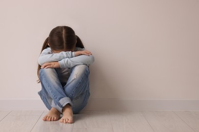 Photo of Child abuse. Upset little girl sitting on floor near light wall indoors, space for text