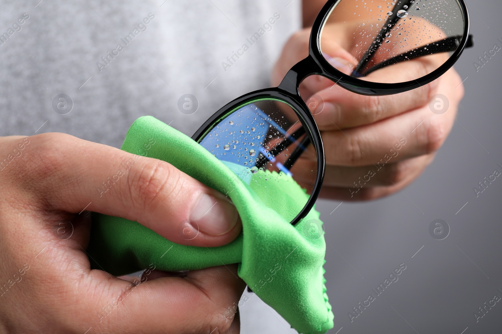 Photo of Man wiping glasses with microfiber cloth on grey background, closeup