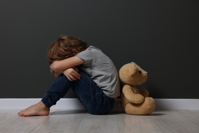 Photo of Child abuse. Upset boy with toy sitting on floor near grey wall