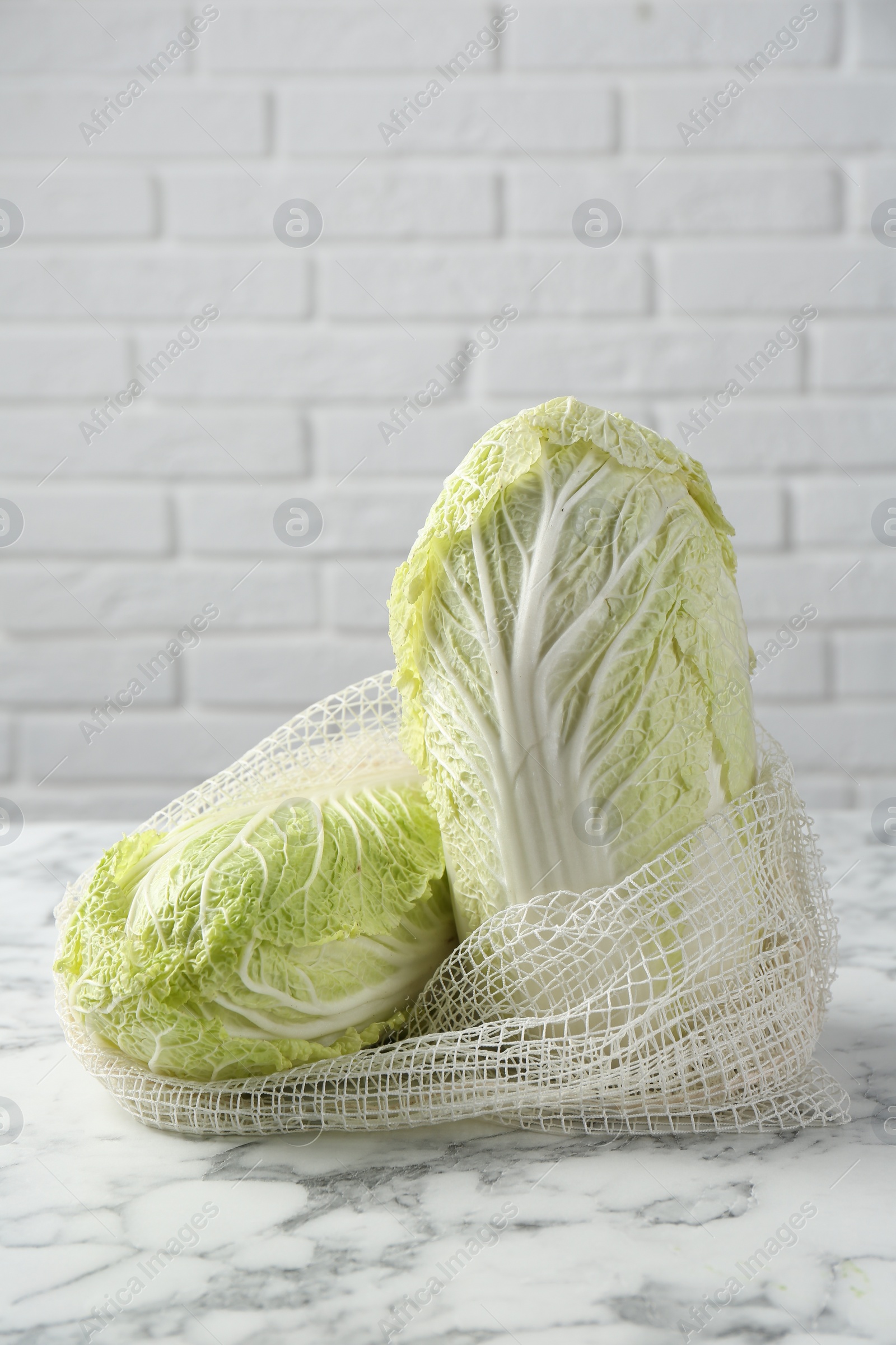 Photo of Fresh Chinese cabbages in string bag on white marble table near brick wall