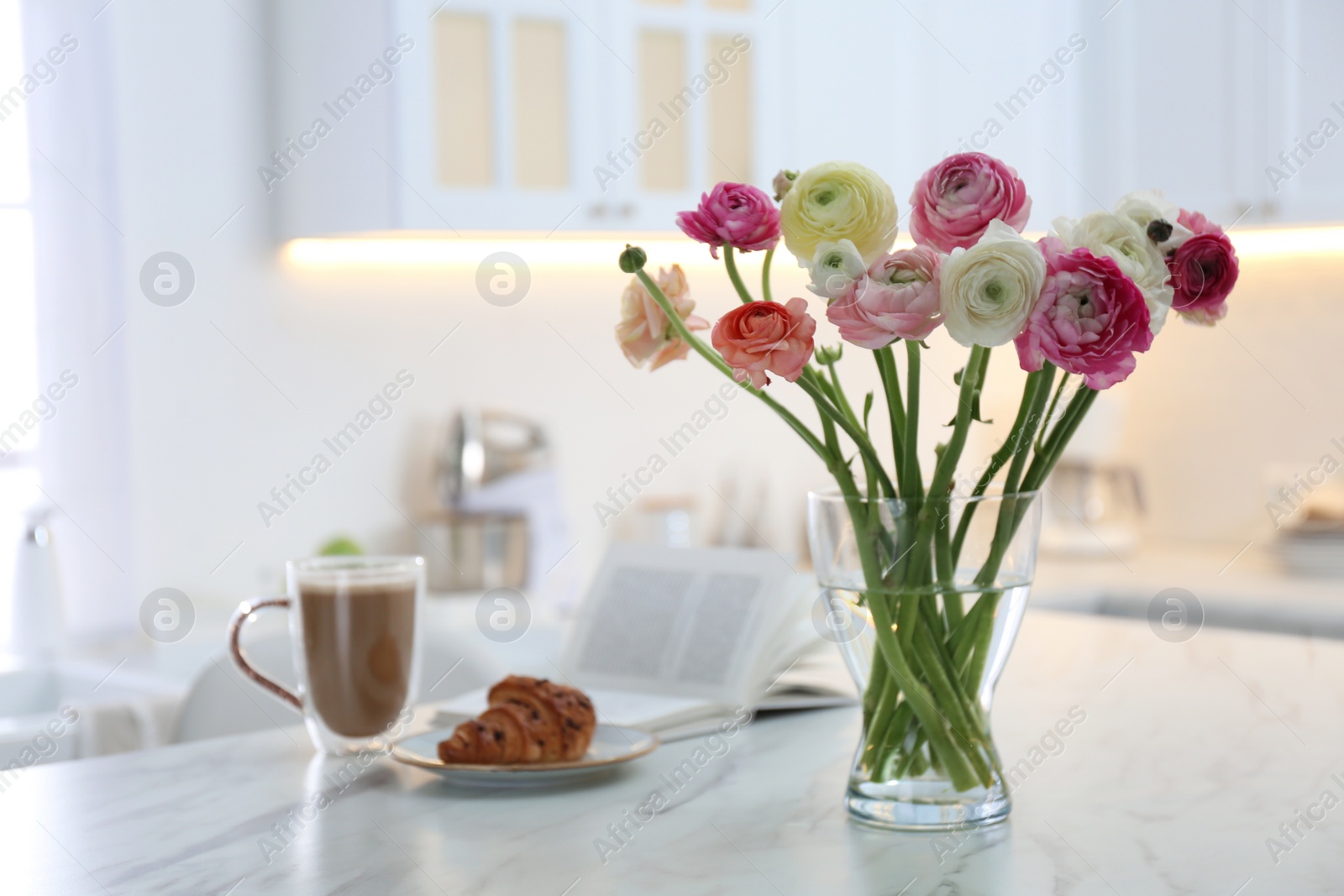 Photo of Beautiful fresh ranunculus flowers near cup of coffee, croissant and book on table in kitchen