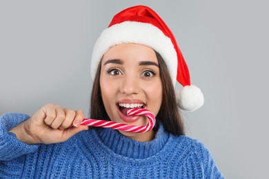 Surprised young woman in blue sweater and Santa hat eating candy cane on grey background. Celebrating Christmas