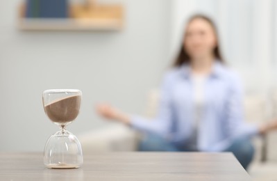 Photo of Hourglass with flowing sand on desk. Woman meditating indoors, selective focus