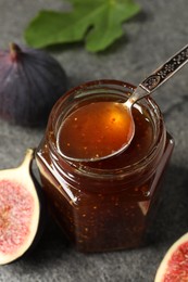 Photo of Glass jar of tasty sweet jam with spoon and fresh figs on grey table, closeup