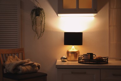 Stylish lamp, cups and croissant on white cabinet in room. Interior element