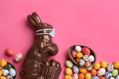 Photo of Chocolate bunny with protective mask and eggs on pink background, flat lay. Easter holiday during COVID-19 quarantine