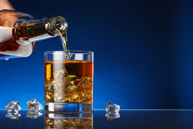 Pouring whiskey into glass with ice cubes at table against dark blue background, space for text