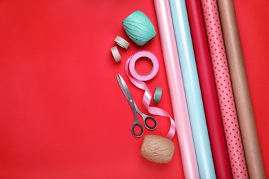 Rolls of wrapping papers, scissors and ribbons on red background, flat lay. Space for text