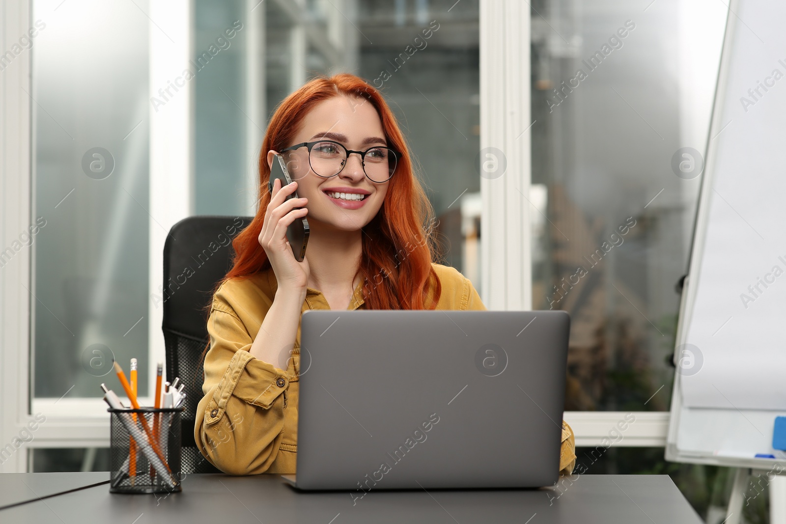 Photo of Happy woman talking on smartphone while working with laptop at desk in office