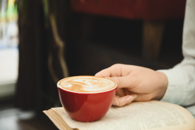 Woman with cup of coffee reading book indoors, closeup