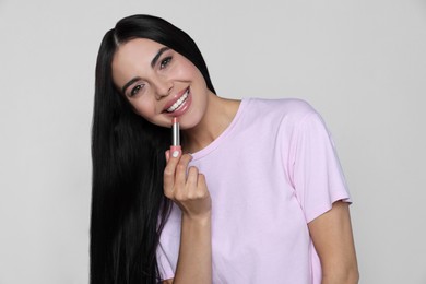 Young woman with beautiful makeup holding nude lipstick on light gray background