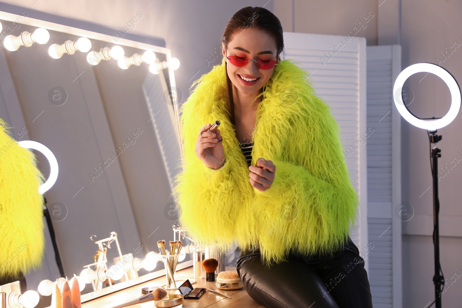 Photo of Young woman applying make up near illuminated mirror indoors