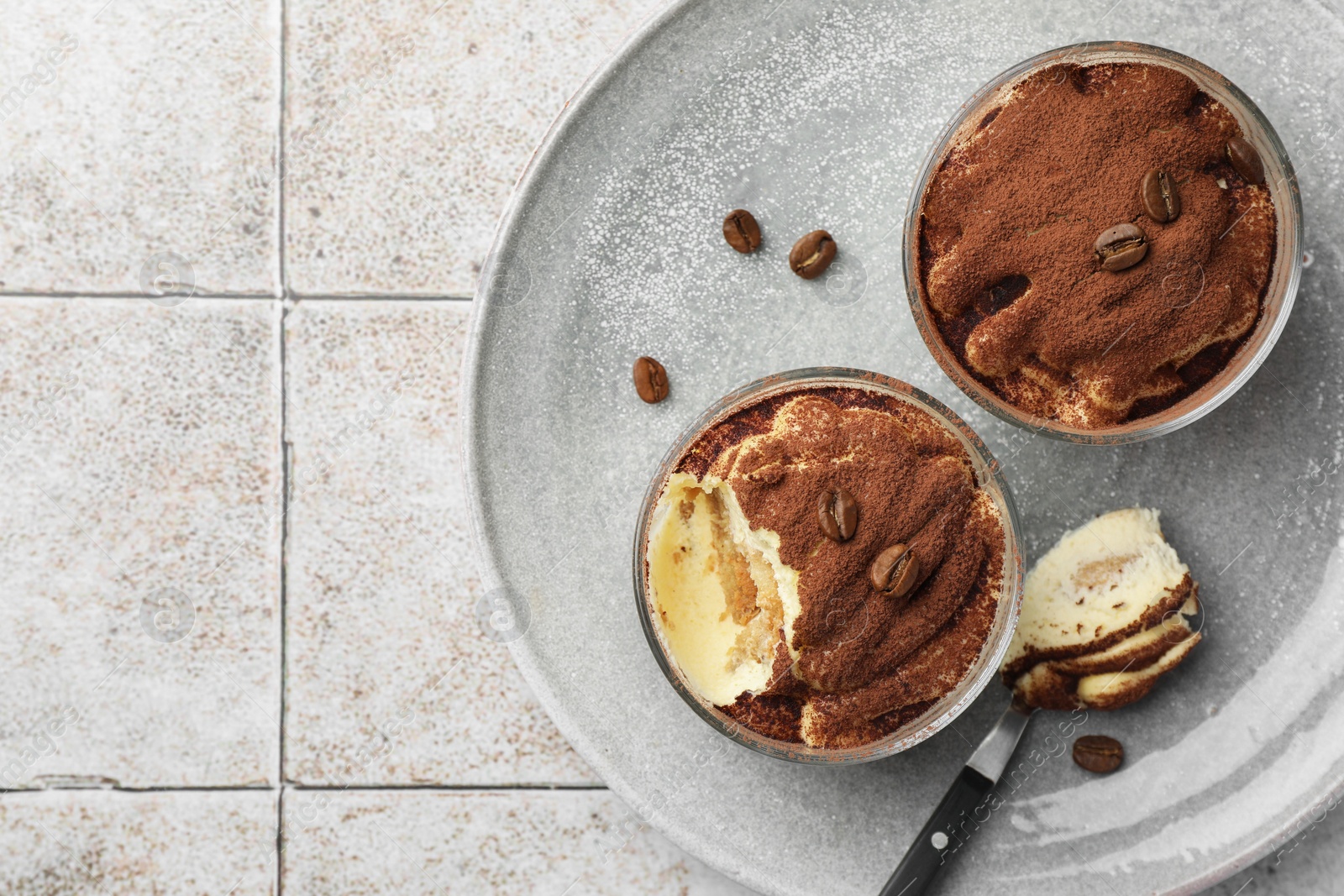 Photo of Delicious tiramisu in glasses, spoon and coffee beans on light tiled table, top view. Space for text