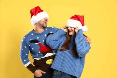 Couple wearing Christmas sweaters and Santa hats on yellow background