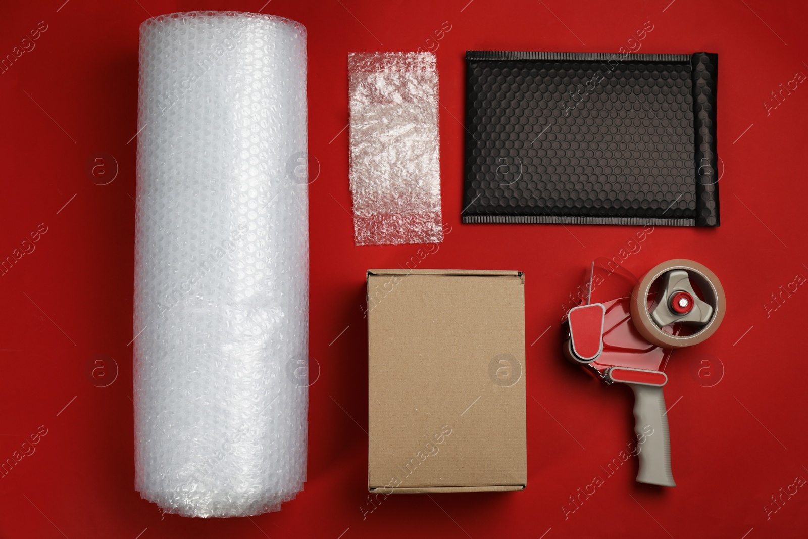 Photo of Bubble wrap with cardboard box, padded envelope and tape dispenser on red background, flat lay