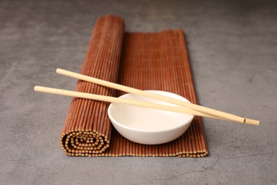 Photo of Rolled bamboo mat, chopsticks and bowl on grey table, closeup