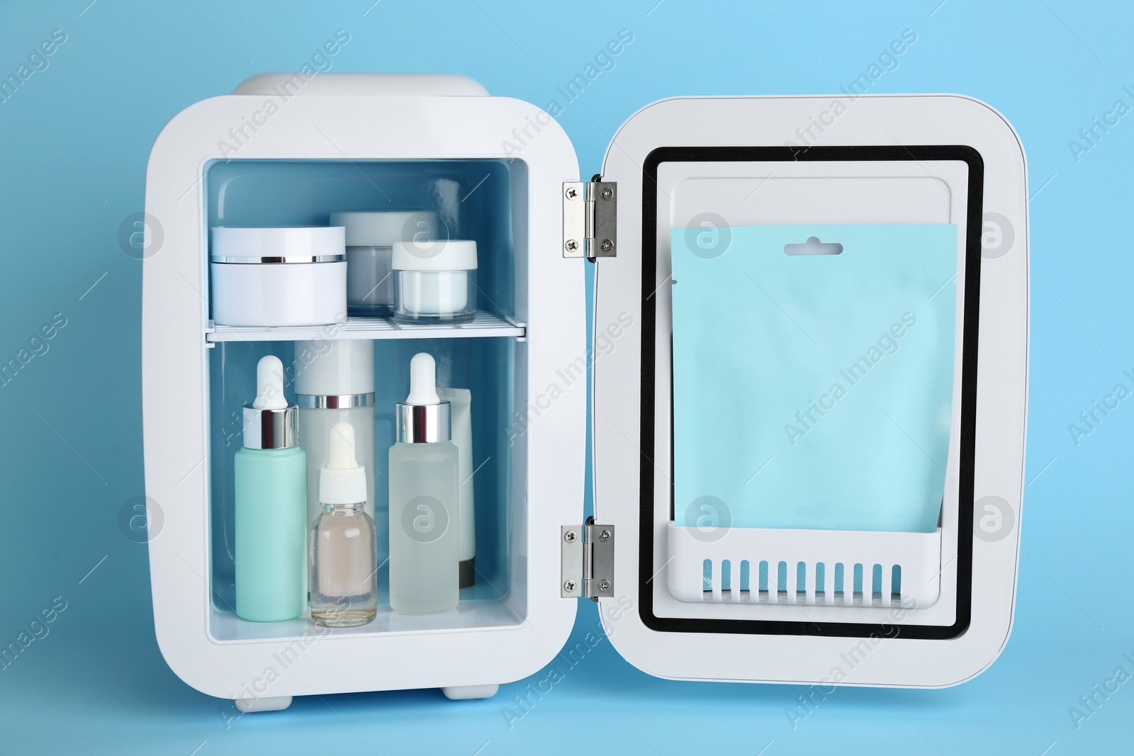 Photo of White cosmetics refrigerator with skin care products on light blue background