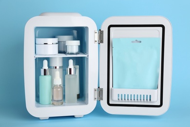 White cosmetics refrigerator with skin care products on light blue background