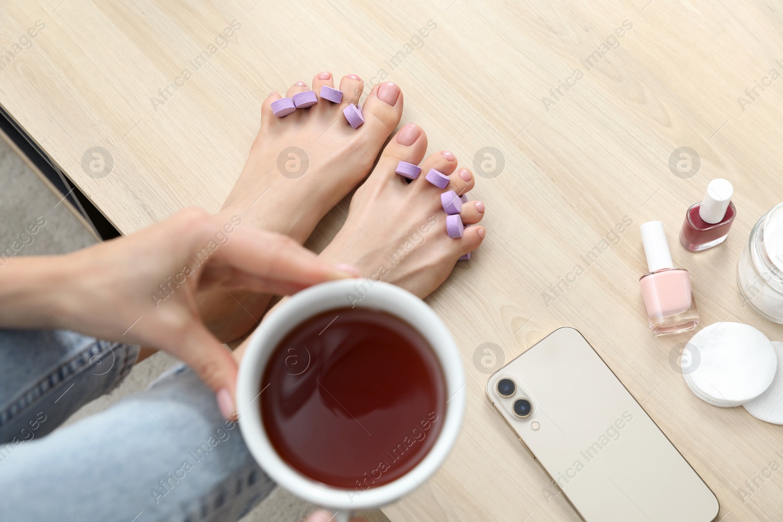 Photo of Woman with cup of tea preparing toenails for pedicure on table at home, closeup