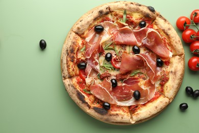 Tasty pizza with cured ham, olives and tomatoes on green background, top view. Space for text