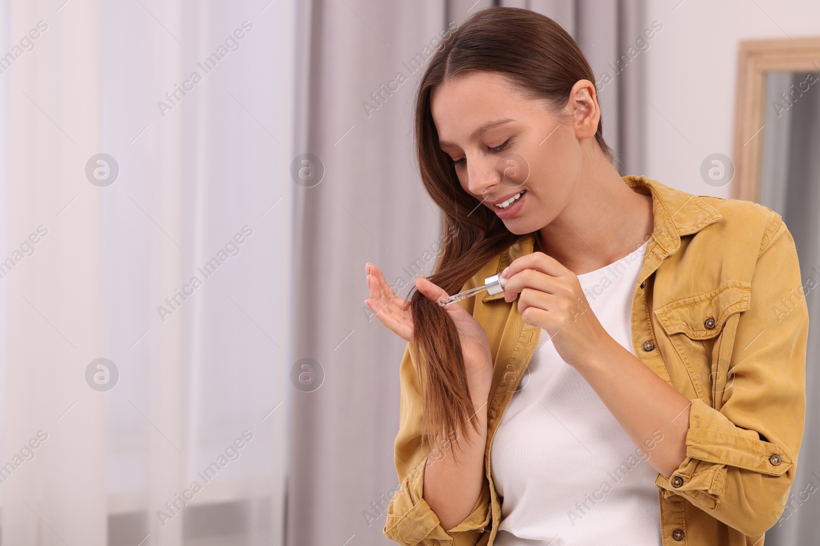 Photo of Beautiful woman applying serum onto hair indoors, space for text