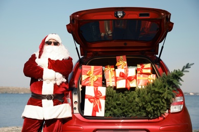 Authentic Santa Claus near car with open trunk full of presents and fir tree outdoors